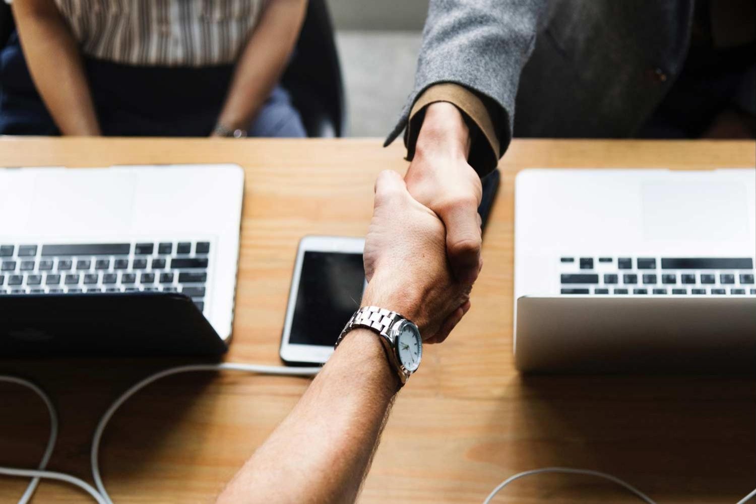Two people shaking hands across a desk between their laptops
